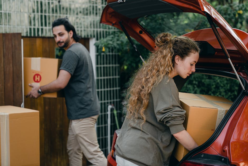A man and woman are engaged in house removal activities outdoors near a residential property, with the woman unloading a large cardboard box from the open trunk of a red hatchback vehicle parked on a driveway. The woman, with long curly hair tied back, is inside the vehicle, carefully placing the box onto the ground while wearing a grey oversized sweatshirt. The man, dressed in a grey t-shirt and beige trousers, is standing nearby holding an additional cardboard box with a printed red 'H' logo, preparing to move it towards a wooden fence that encloses the garden area. Several other cardboard boxes are situated on the driveway and ground, some partially stacked, all wrapped with packing tape to secure their contents, indicating a packing stage of the home relocation. Visible elements include the vehicle's open trunk, motionless in a loading process, a pavement, fencing, greenery in the background, and soft, natural daylight illuminating the scene, which reflects typical house moving preparations facilitated by Catford Man and Van, specialists in removals and furniture transport.
