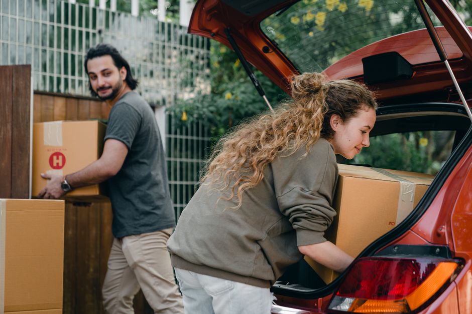 A young woman with long, curly hair wearing a grey sweatshirt is loading a large cardboard box into the open trunk of a red hatchback vehicle parked on a paved driveway, with her head slightly bowed and a slight smile. Behind her, a young man with dark hair and a beard, dressed in a grey t-shirt and beige trousers, is holding another medium-sized cardboard box, preparing to place it on a wooden platform or pile marked with red symbols near a wooden fence. The background features greenery and trees, indicating an outdoor residential setting. Visible packing materials include cardboard boxes, some taped and stacked, and protective wrapping on the furniture items. The scene captures a moment in a home relocation or furniture transport process, with the individuals engaged in loading and organizing household items for moving, with a focus on careful handling and packing which are key aspects of professional removals services, as offered by Catford Man and Van.
