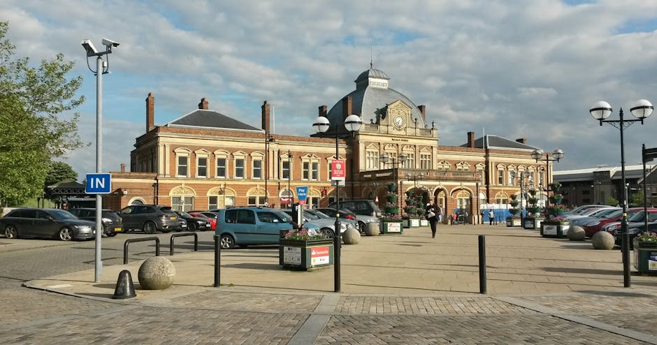 A large, historic brick building with a central clock tower and ornate architectural details, situated behind a spacious parking area with multiple parked cars on either side. The parking lot is lined with black metal bollards, decorative street lamps with double globes, and flower planters, with a blue 'IN' sign indicating traffic flow. There are a few pedestrians walking across the pavement and entering the building. The scene is illuminated by daylight, with a partly cloudy sky overhead and some greenery visible from trees on the left. The parking area appears to be used for visitors or residents, and the building could be a town hall, train station, or similar public structure. The image may relate to house removals or moving services in Catford, highlighting logistical considerations such as parking access for loading and unloading furniture during home relocation processes, as exemplified by the services of Catford Man and Van.