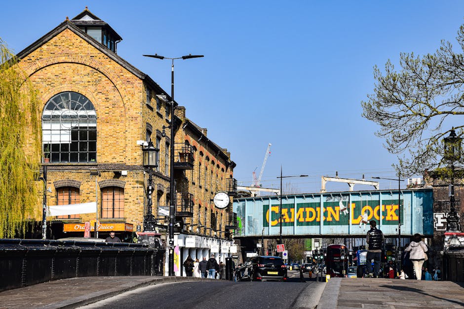 A wide street scene in Catford, London, showing a brick building with large arched and rectangular windows, some covered with wooden boards, and exterior metal fire escapes. On the street, several pedestrians walk along the pavement, and parked cars are visible on both sides. To the right, a painted metal railway bridge with the words 'Camden Lock' spans the road. In the background, construction cranes and other buildings are visible under a bright blue sky, with a few trees on the left and right edges. Streetlights and a clock are mounted on black poles along the sidewalk, indicating it is daytime. This scene captures an everyday view in Catford, suitable for illustrating house removals, moving logistics, or furniture transport in a professional context, as facilitated by Catford Man and Van.