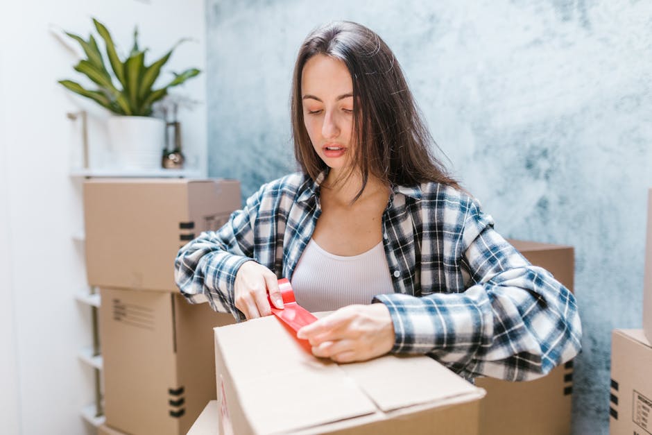 A young woman with long dark hair, wearing a blue and white checked shirt over a white top, is standing indoors in a room with a blue textured wall and a white shelf with a potted plant. She is in the process of sealing a cardboard box with red tape, holding the tape dispenser in her right hand and pressing the tape onto the box with her left hand. Several packed cardboard boxes of various sizes, some with printed labels and black tape, are stacked behind her, indicating a home relocation or moving process. The environment is well-lit, and the scene captures the packing stage of furniture transport or home moving assistance, demonstrating the packing and organizing aspects of residential removals, as performed by a company like Catford Man and Van.